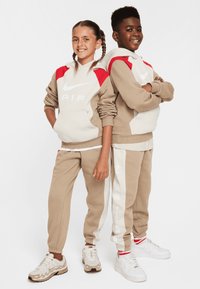 Dvaja chlapci majú na sebe zladené oblečenie Nike. Mikiny sú béžové s červenými akcentmi, zatiaľ čo tepláky sú béžové s bielymi prúžkami a logami.