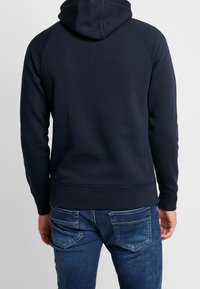 Sudadera azul marino con un bolsillo tipo canguro, puños y bajo acanalados, y capucha forrada. Lleva puesta con jeans de mezclilla azul claro con bolsillos traseros.