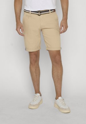 Homme portant un short beige retroussé, une ceinture blanche avec des rayures bleues et marron, des baskets blanches et un T-shirt blanc rentré.