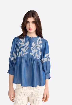 Chemisier en denim bleu avec des manches bouffantes trois-quarts, un devant boutonné et des motifs de feuilles brodés en blanc sur la poitrine et les épaules.