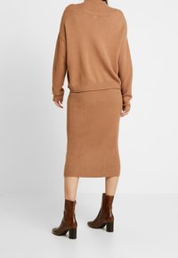 Maglione lavorato a maglia color sabbia con colletto alto e texture a coste, abbinato a una gonna a matita a coste coordinata. Stivali alla caviglia marroni completano l'outfit.