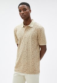 Polo de manga corta en beige con un patrón floral, con cuello y botones, hecho de una tela suave y texturizada.