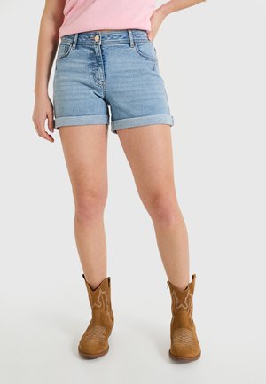 Pantalones cortos de mezclilla en azul claro con puños enrollados, con bolsillos frontales y cierre de botón de metal, usados con botas de vaquero marrones.