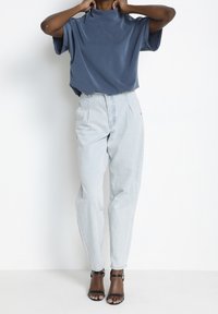 Lös passform blå topp med hög halsringning, kombinerad med ljusa denim byxor med hög midja och veck samt svarta sandaler med remmar.