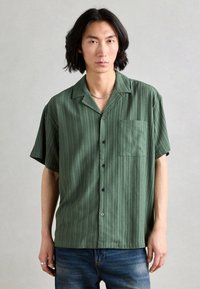 Button-up short-sleeve shirt in dark green striped fabric, featuring a patch pocket and black buttons. Model paired with blue jeans.