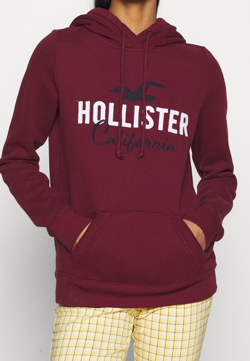 Felpa con cappuccio color bordeaux con tasca a marsupio frontale, cappuccio con coulisse e scritta bianca "HOLLISTER California". Tessuto morbido e strutturato.