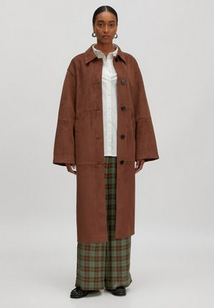 Brauner Wildleder-Mantel mit kragenförmigem Ausschnitt, oversized Passform, schwarzen Knöpfen und großen Ärmeln, getragen über einem karierter Unterteil.