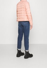 Giacca a vento rosa chiaro con cuciture orizzontali, abbinata a jeans blu scuro e stivali neri alla caviglia. La giacca presenta un collo alto.