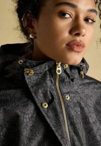 Čierna vodoodolná bunda s kvetinovým uzorom a vysokým golierom, zlatým zipsom a kovovými akcentmi. Obsahuje nastaviteľnú šnúrku okolo kapucne.