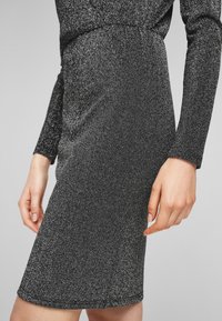 Svart glittrig klänning med långa ärmar, figurnära design och slät textur. Sjöna slutar strax ovanför knäet och har en diskret glans.