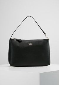 Sac à main Guess en cuir noir avec une bandoulière unique, une fermeture éclair et une plaque de logo argentée à l'avant, présenté sur un socle blanc.