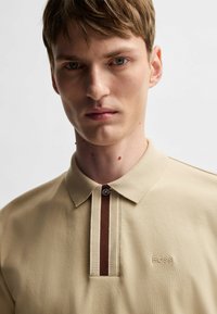Beige polo shirt with a collared neckline, featuring a brown vertical stripe and an embroidered "BOSS" logo on the chest. Textured fabric.
