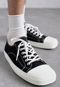Sneaker nere in tela con punta in gomma bianca, lacci bianchi e cuciture a contrasto. Indossati con calzini bianchi sopra la caviglia.