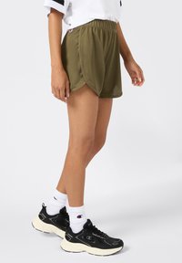 Pantaloni da atletica in rete verde oliva con orlo curvo, vita elastica e accento con logo. Abbinati a sneakers nere e calze bianche a crew.