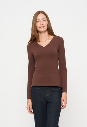 Femme aux cheveux longs et raides portant un haut marron ajusté à manches longues et un jean bleu foncé, debout devant un fond blanc uni.