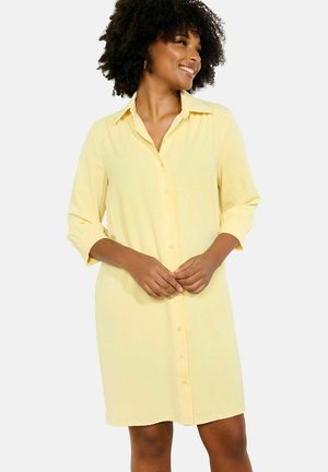 Woman with curly hair smiling, wearing a long-sleeve, buttoned yellow shirt dress, standing with hands loosely clasped.