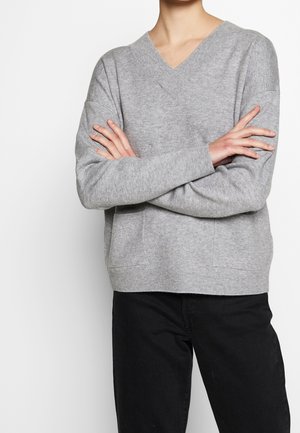 Person mit verschränkten Armen, trägt einen hellgrauen V-Ausschnitt-Pullover und schwarze Hose vor schlichtem weißem Hintergrund.