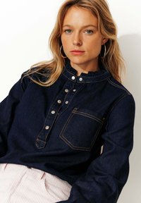 Chemise en denim avec un col volanté, des boutons sur le devant, des coutures décoratives et une poche au niveau de la poitrine. Porte avec un pantalon en velours côtelé clair.