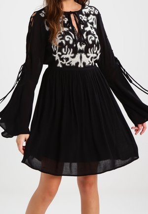 Robe noire longueur genou avec motifs floraux brodés blancs sur la poitrine et les épaules, manches longues évasées avec détails noués, et taille froncée.