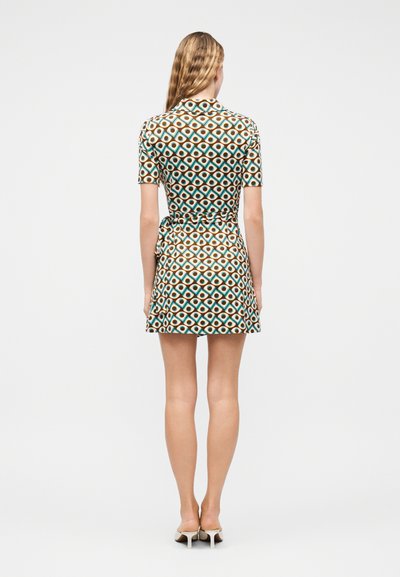 Γυναίκα με μακριά μαλλιά που φοράει ένα κοντομάνικο κοντό φόρεμα με γεωμετρικό μοτίβο και λευκά παπούτσια με τακούνι, που στέκεται γυρισμένη προς τα πίσω.