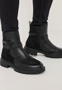 Bottines noires en cuir lisse et en daim, avec une fermeture éclair sur le côté, des accents de boucle et une semelle en caoutchouc épaisse avec des crampons.