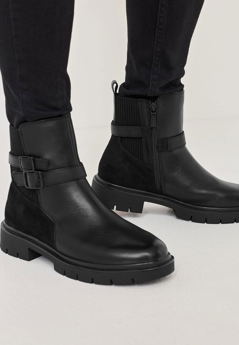 Bottines noires en cuir lisse et en daim, avec une fermeture éclair sur le côté, des accents de boucle et une semelle en caoutchouc épaisse avec des crampons.