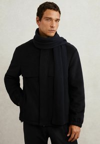 Chaqueta de lana negra con cremallera en la parte delantera, dos bolsillos en el pecho y puños de canalé. Envueltos en una bufanda de canalé negra a juego. Textura suave.