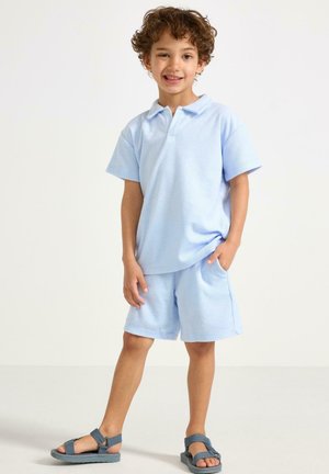 Lyseblå polo- og shorts-sæt i frotté, med krave, korte ærmer og sidelommer. Kombineret med grå sandaler.