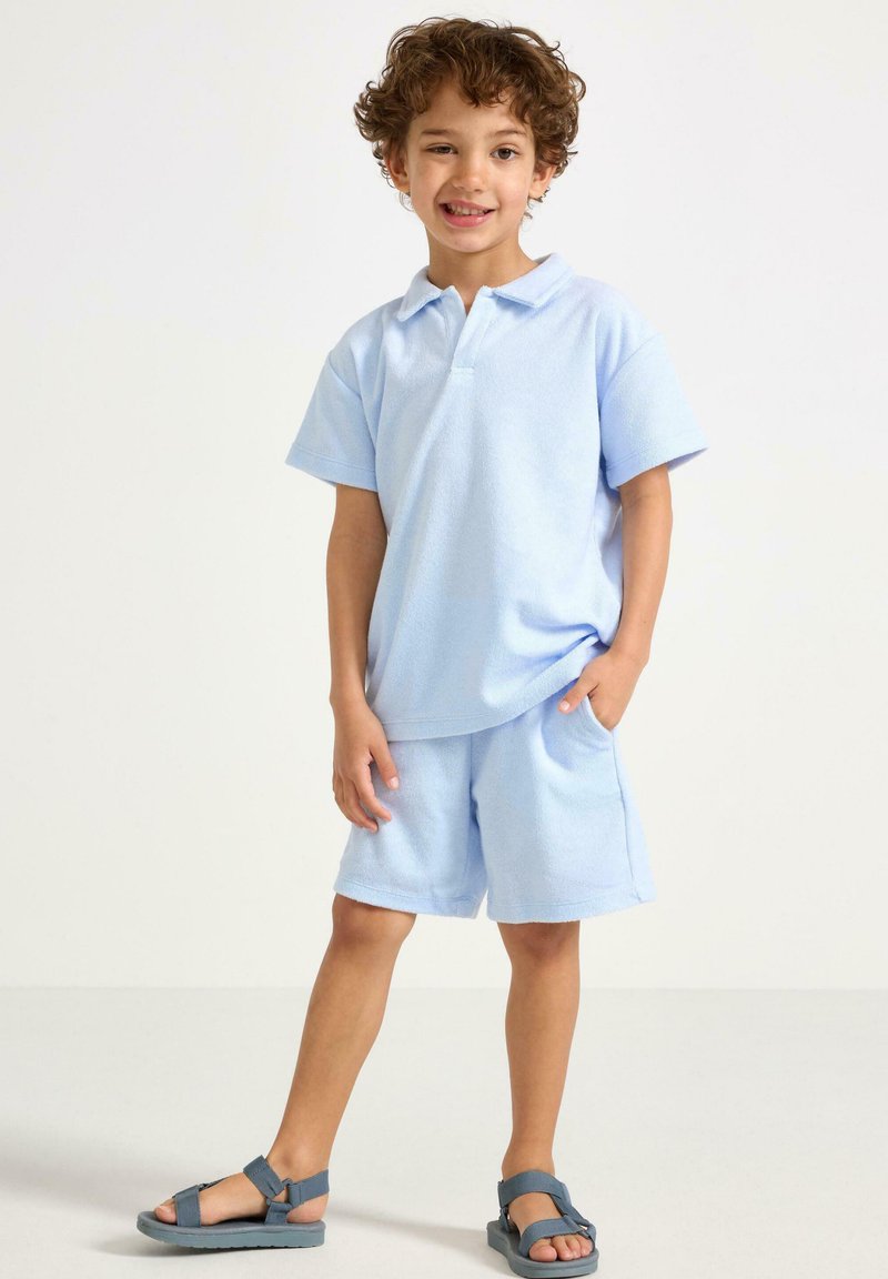 Lyseblå polo- og shorts-sæt i frotté, med krave, korte ærmer og sidelommer. Kombineret med grå sandaler.