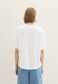 T-shirt bianco oversize con scollatura tonda, maniche corte e orlo dritto. Indossato sopra pantaloni larghi blu chiaro. Tessuto dalla consistenza liscia.
