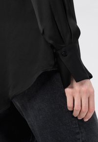 Svart sjevtockat långärmad skjorta med knäppt manschett, slät textur och böjd nederkant, i kombination med mörk denimjeans.