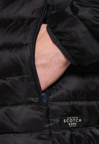 Hand in schwarzer Daunenjackentasche mit Reißverschluss neben einem Etikett mit der Aufschrift "Scotch & Soda Hand Crafted with Love."