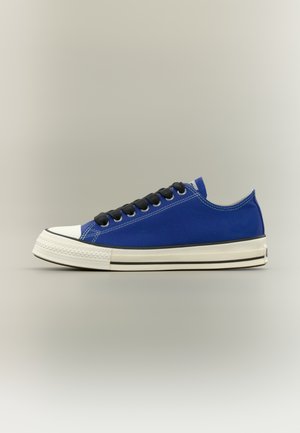 Modri platneni nizki supergi z črnimi vezalkami, belim gumijastim ščitnikom na prstih in belim podplatom s črnimi črtami na nevtralnem ozadju.