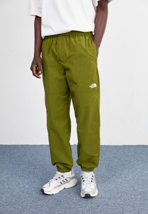 Osoba ubrana w oliwkowe spodnie dresowe The North Face i białe sneakersy, stojąca na szarym dywanie z jedną ręką w kieszeni.