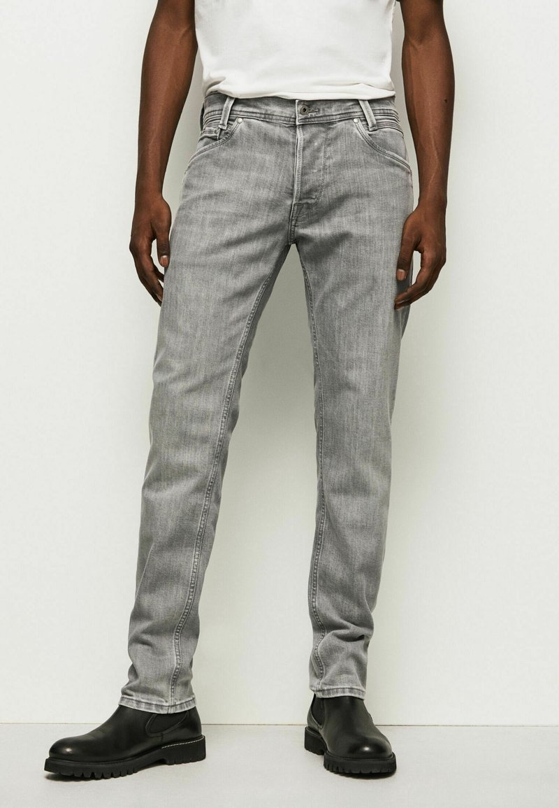 Homme portant un jean slim gris clair, des bottes en cuir noir et une chemise blanche, debout devant un fond clair uni.