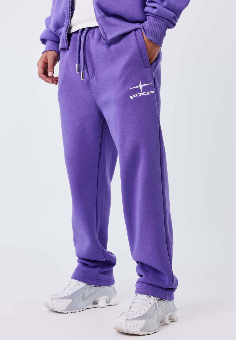 Persona vestida con pantalones de chándal morados PXP y una sudadera con cremallera a juego, con zapatillas blancas sobre un fondo blanco.