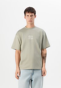 Ung mand med kort brunt hår iført en lys grå oversized t-shirt med hvidt logo og lys blå jeans, står foran en hvid baggrund.