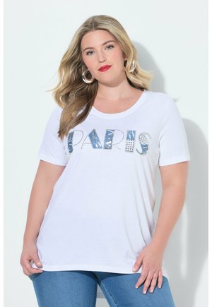 Kvinde med langt blondt hår iført en hvid T-shirt med dekoreret "PARIS"-tekst og blå jeans står foran en ensfarvet lys baggrund.