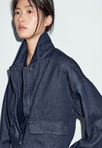 Giovane donna con capelli scuri raccolti, indossa una giacca di denim scuro con colletto alto e dettagli con bottoni su uno sfondo neutro.