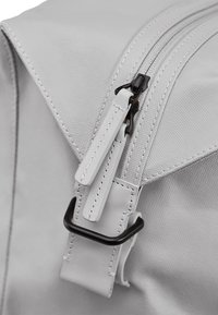 Sac à dos en tissu gris avec une surface texturée, mettant en avant une fermeture éclair bien visible, des accents en cuir et un crochet en métal noir.