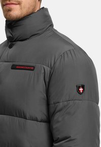 Man met donkergrijze gewatteerde jas met hoge kraag, rode "KRONSTADT"-logo op de borst en schildembleem op de bovenarm.