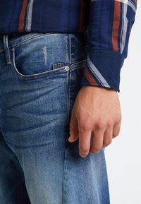 Mano masculina junto a un bolsillo de unos vaqueros azules desgastados y una camisa azul de manga larga con rayas rojas y blancas en el puño.
