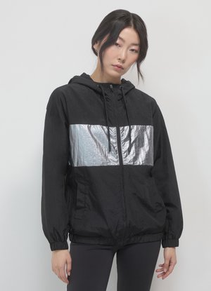 Una donna indossa una giacca nera con cappuccio, munita di una larga striscia riflettente argentea sul petto, abbinata a leggings scuri su uno sfondo semplice.