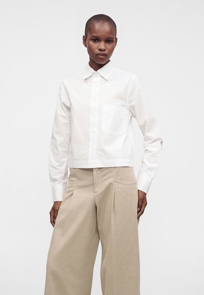 Personne portant une chemise blanche à boutons avec une poche avant et un pantalon beige à jambes larges, debout devant un fond clair uni.
