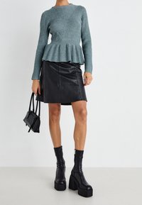 Pull turquoise côtelé à taille peplum, associé à une mini-jupe en cuir noire. Le modèle porte des bottes noires montantes à plateforme et tient un petit sac.