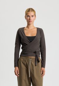 Elegancka bluzka w ciemnym brązowym kolorze z długimi rękawami i głębokim dekoltem w kształcie litery V, z dodatkowym wiązaniem w talii. Nosi się ją na czarnej koszulce na ramiączkach i zestawia z luźnymi, khaki spodniami.