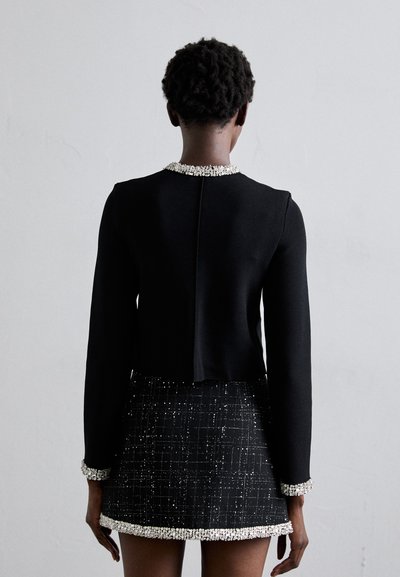 Svart kort jacka med en krage prydd med juveler, matchad med en svart minikjol med ett texturerat mönster och dekorerad fåll.