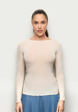 Kobieta o ciemnych włosach spiętych do tyłu, ubrana w dopasowaną kremową bluzkę z długim rękawem i niebieską spódnicę, stojąca na jednolitym jasnym tle.