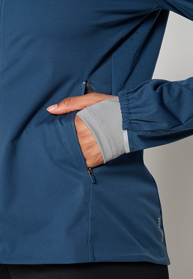 Marineblaue Softshell-Jacke mit einer Reißverschlusstasche, kontrastierenden grauen Saumdetails und atmungsaktiven Perforationen am Ärmel.