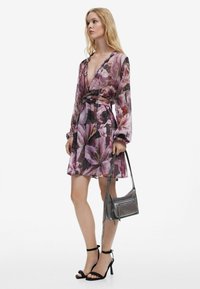 Blumiges Mini-Kleid mit langen, durchsichtigen Ärmeln, tiefem V-Ausschnitt und tailliertem Bund. Ausgestattet mit rosa und lila Farbtönen, kombiniert mit einer grauen Handtasche und schwarzen Absätzen.